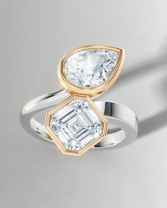A split-band ring featuring a pear-cut clear gemstone in a gold bezel on one side and an emerald-cut clear gemstone in a silver bezel on the other, set against a neutral gray background.