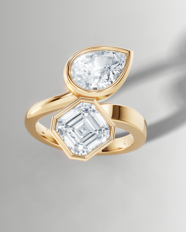 A gold ring featuring two clear gemstones, one pear-cut and one emerald-cut, set against a neutral gray background with a soft shadow.