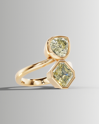A gold ring featuring two light green gemstones, one round and one hexagonal, set on a smooth gray gradient background.