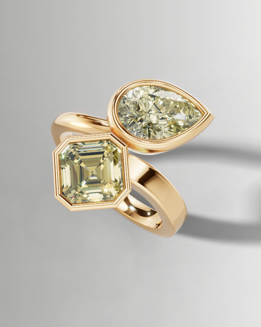 A gold ring featuring a large emerald-cut pale yellow gemstone and a smaller pear-cut pale yellow gemstone, displayed on a clean light gray background with a soft shadow.