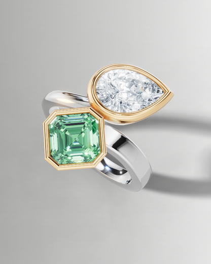 A two-stone ring featuring a square emerald-cut green gemstone in a gold setting and a pear-cut clear gemstone in a gold setting, mounted on a split silver and gold band, shown on a light gray background.