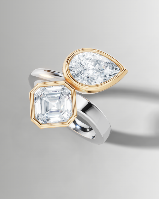 A split-design ring featuring a large emerald-cut clear gemstone in a gold bezel setting and a pear-cut clear gemstone in a silver bezel setting, mounted on a polished silver band.