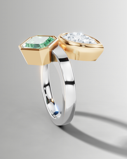 A sculptural two-stone ring featuring a yellow gold hexagonal setting with a green emerald-cut gemstone and a silver-toned band with a clear round-cut gemstone, shown on a neutral gray background.
