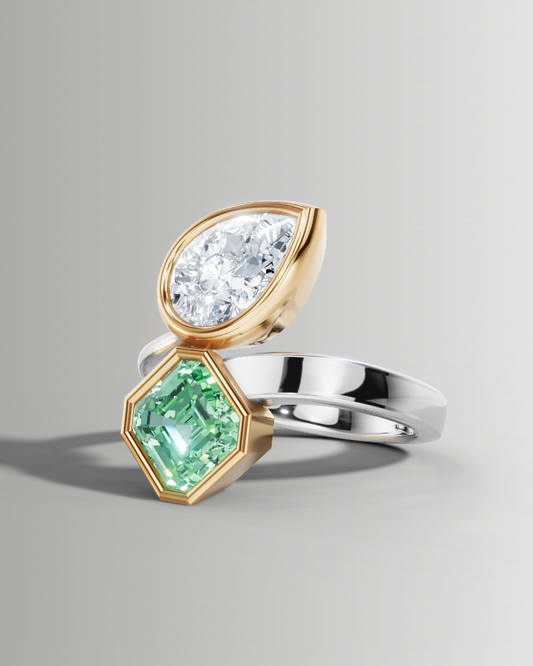A two-stone ring featuring a pear-cut clear gemstone in a gold setting and a hexagonal green gemstone in a yellow-gold setting, mounted on a silver-colored band, shown on a neutral gray background.