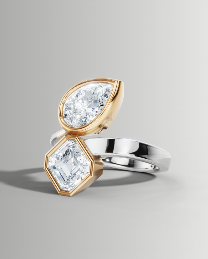 A two-stone diamond ring featuring one pear-cut diamond in a gold bezel setting and one emerald-cut diamond in a silver bezel setting, mounted on a split silver and gold band, shown on a neutral gray background.